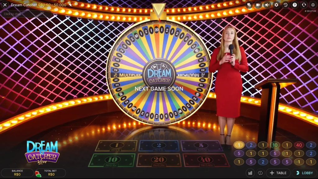 Uma interface de jogo de cassino ao vivo mostrando uma roda "Dream Catcher" com segmentos multicoloridos e números. Uma apresentadora em um vestido vermelho está ao lado da roda, segurando um microfone. A interface exibe opções de apostas e controles do jogo.