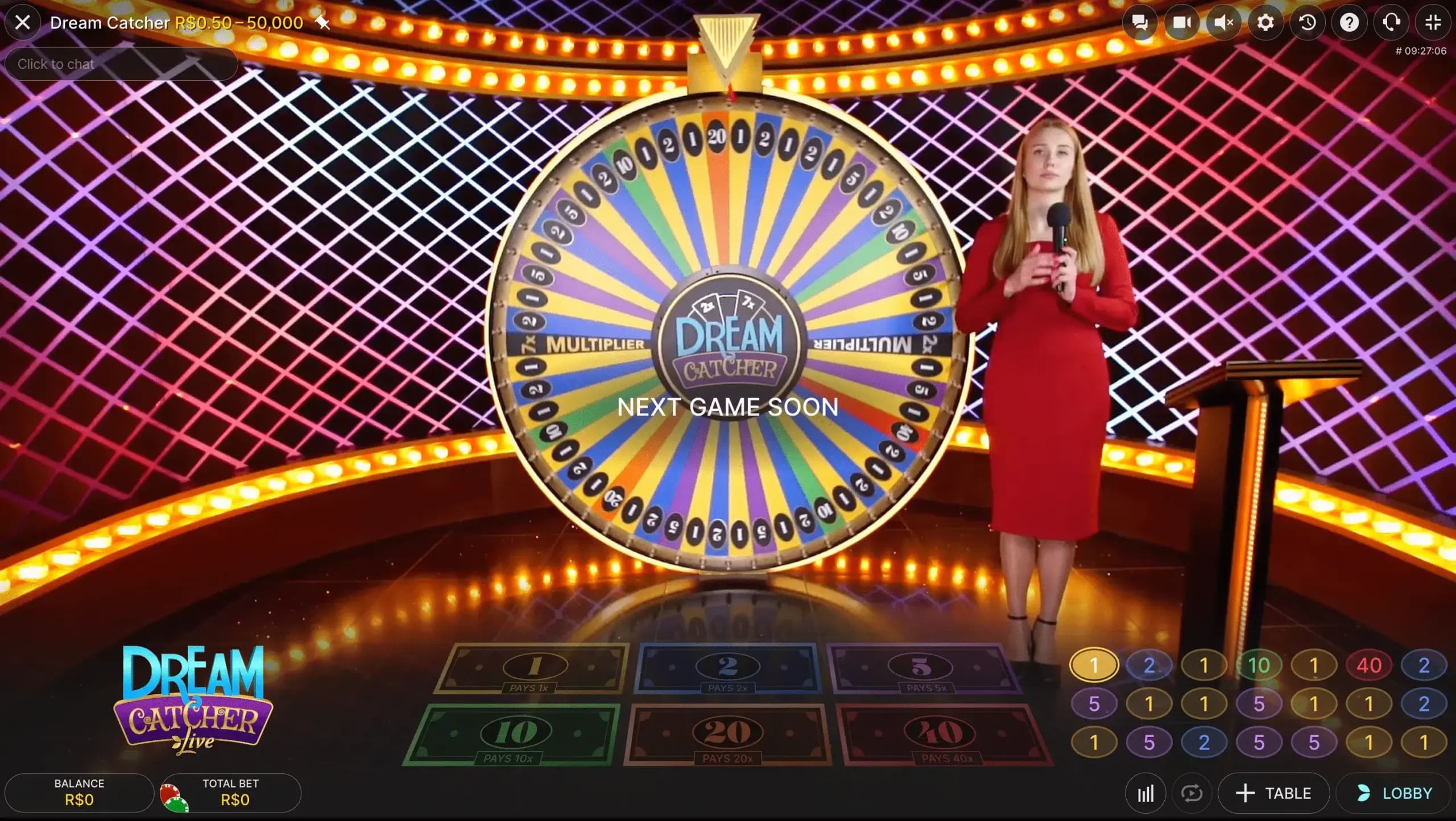 Un juego de casino en vivo ambientado con una gran y colorida rueda "Dream Catcher". La rueda tiene múltiples segmentos numerados en varios colores. Una presentadora con un vestido rojo está de pie junto a la rueda, sosteniendo un micrófono. El fondo tiene un patrón vibrante de luces rosas y moradas. Debajo de la rueda hay una interfaz de apuestas con opciones para diferentes apuestas numéricas.