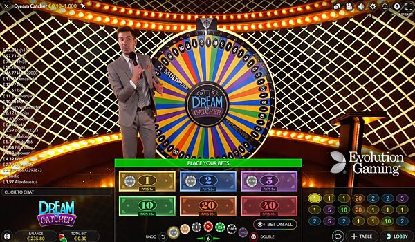 Otra vista del juego de casino en vivo "Dream Catcher". Un presentador con traje gris se encuentra junto a la gran rueda colorida. El set tiene un fondo de celosía dorada con luces de cadena. Las opciones de apuesta se muestran en la parte inferior de la pantalla, junto con el logotipo del juego y la marca Evolution Gaming.