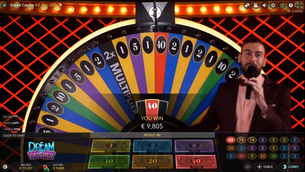 Viene mostrato un game show del casinò dal vivo chiamato "Dream Catcher". La caratteristica principale è una grande ruota colorata divisa in segmenti con numeri e moltiplicatori diversi. La ruota si è fermata sul numero 40, indicato da "HAI VINTO €9.805" al centro. Un presentatore in abito nero e papillon è visibile sulla destra, che parla in un microfono. Lo sfondo ha un motivo incrociato rosso con luci brillanti. Nella parte inferiore dello schermo ci sono le opzioni di scommessa per diversi numeri e moltiplicatori. Il saldo del giocatore è mostrato come €19.257.