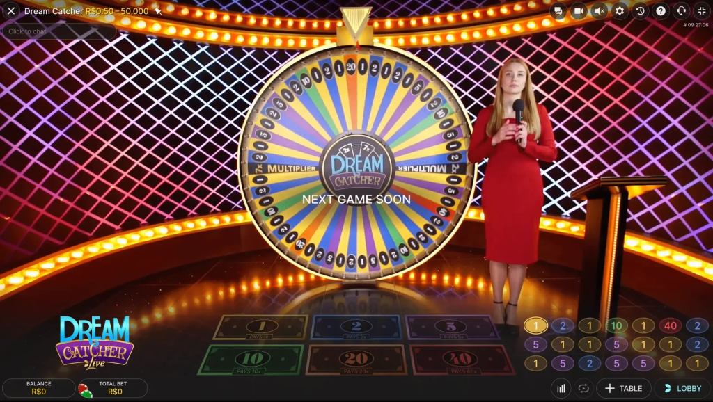 Una donna in un abito rosso si trova accanto a una grande ruota colorata che gira etichettata "Dream Catcher" in un ambiente da game show. La ruota presenta numeri e moltiplicatori, con sezioni per 1, 2, 5, 10, 20 e 40. Lo sfondo è un motivo a griglia illuminato e il testo "NEXT GAME SOON" è visualizzato sulla ruota. Nella parte inferiore dell'immagine, ci sono indicatori per gli importi del saldo e della scommessa totale.