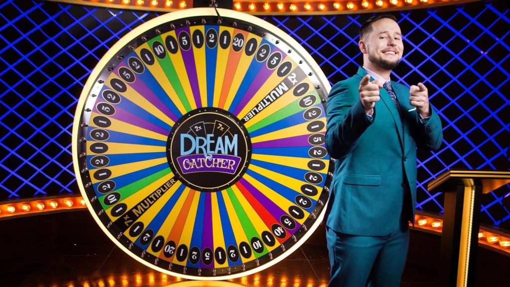 A game show set featuring a large colorful wheel labeled "Dream Catcher" with numbers and multipliers. A smiling host in a teal suit stands next to the wheel, gesturing towards it.