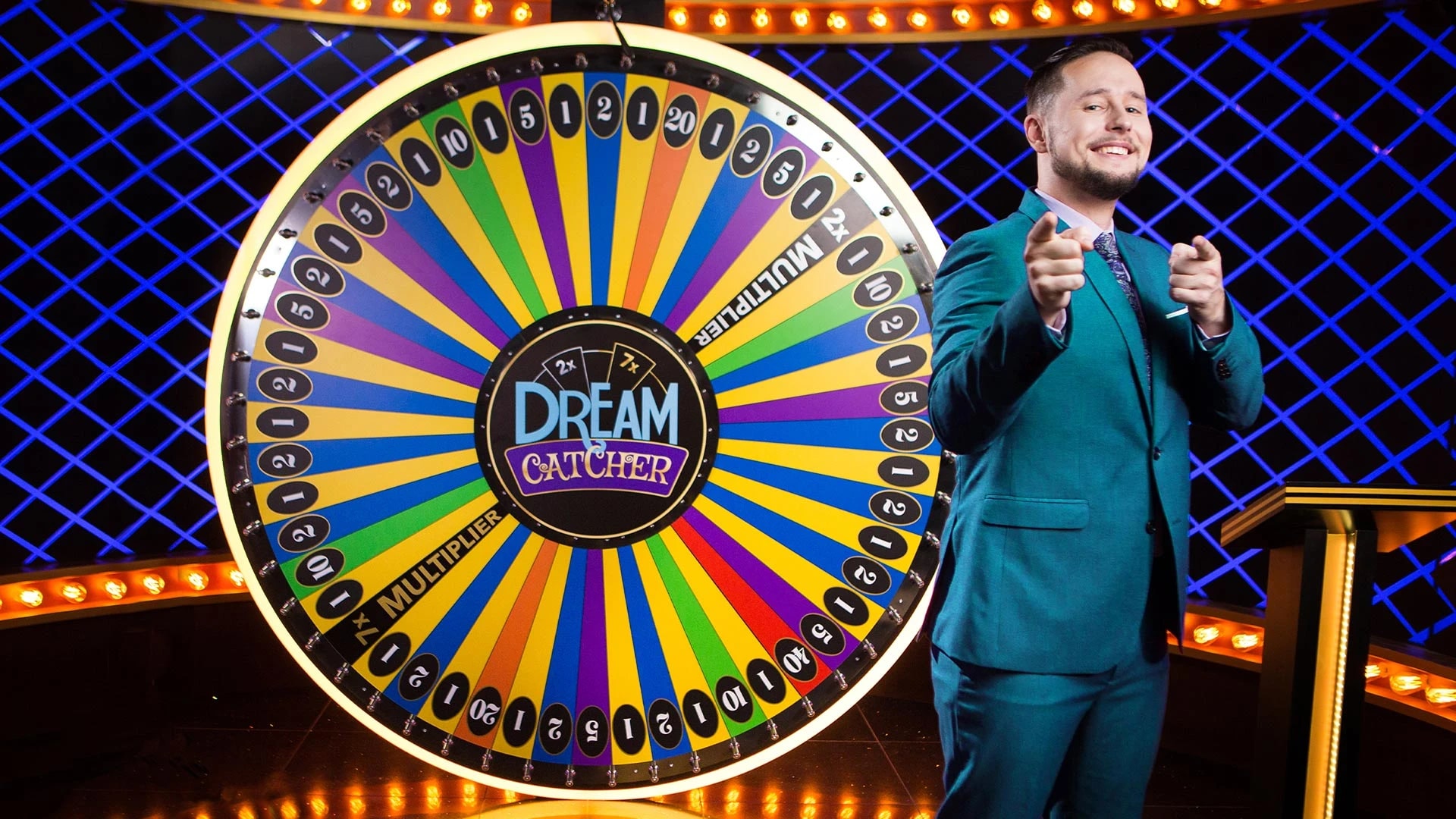 A colorful Dream Catcher wheel with numbered segments and multipliers, next to a smiling game host in a teal suit giving thumbs up. The wheel features vibrant rainbow colors and is set against a blue-lit studio backdrop with illuminated borders.